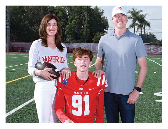 Three people stand on a football field with one person in a red football uniform holding a football while the other two stand on either side, smiling. Description generated by AI