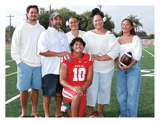 A group of six people, five standing and one sitting in front wearing a red sports jersey, are smiling outdoors on a field with trees and power lines in the background. Description generated by AI