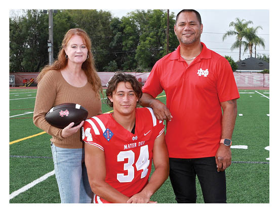 Three people are posing on a football field with one person in a red football uniform holding a football and the other two standing beside them, one wearing a brown sweater and the other a red polo shirt. Description generated by AI