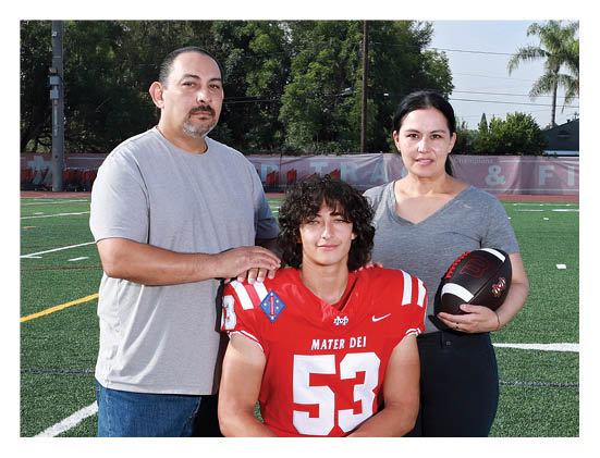 A person in a red football uniform with the number 53 is kneeling on a football field while two adults stand behind them, one resting a hand on their shoulder and the other holding a football. Description generated by AI