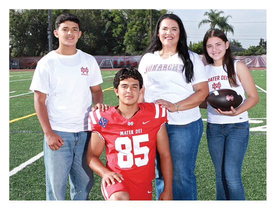 Four people stand on a sports field with one person wearing a red football uniform and the others wearing casual white shirts, with one holding a football. Description generated by AI