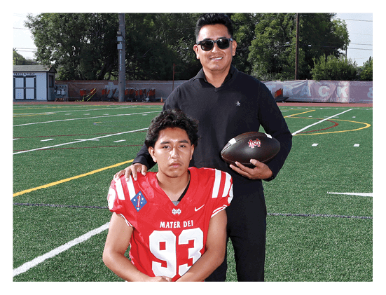 A person in a red football jersey with the number 93 sits on a field while another person stands behind them wearing sunglasses and holding a football. Description generated by AI