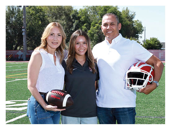 Three adults stand closely together on a football field with one holding a football and another holding a football helmet, all smiling and dressed casually. Description generated by AI