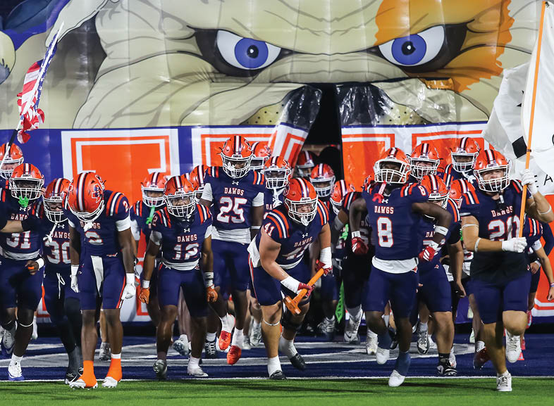 Four people wearing black football uniforms with “VISTA" on the front are standing together in a stadium with a crowd in the background, two of them holding helmets raised above their heads. Description generated by AI