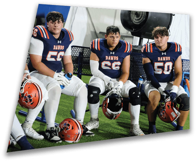 Four people in dark football uniforms and helmets stand in a row on a field, appearing focused and ready for action. Description generated by AI
