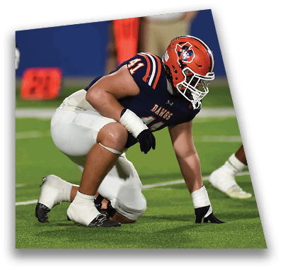 A person in football gear and helmet is crouched on a field with other players lined up in the background during a game. Description generated by AI