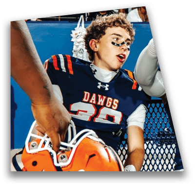 A person in football gear and helmet is crouched on a field with other players lined up in the background during a game. Description generated by AI
