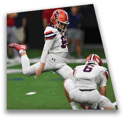 A person in football gear and helmet is crouched on a field with other players lined up in the background during a game. Description generated by AI