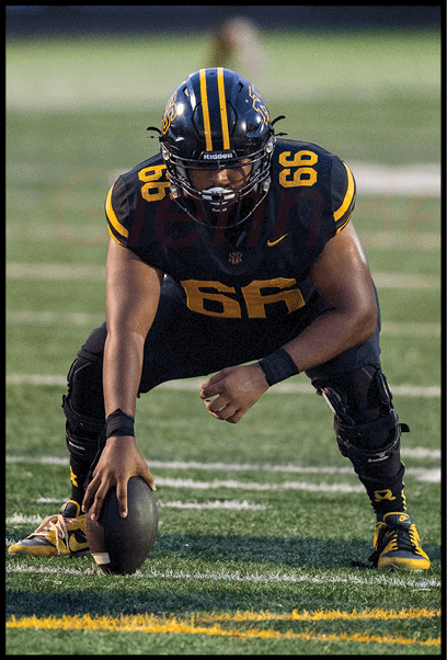 A person in a black and yellow football uniform is crouched on a field holding a football, preparing to snap it, with other players visible nearby. Description generated by AI