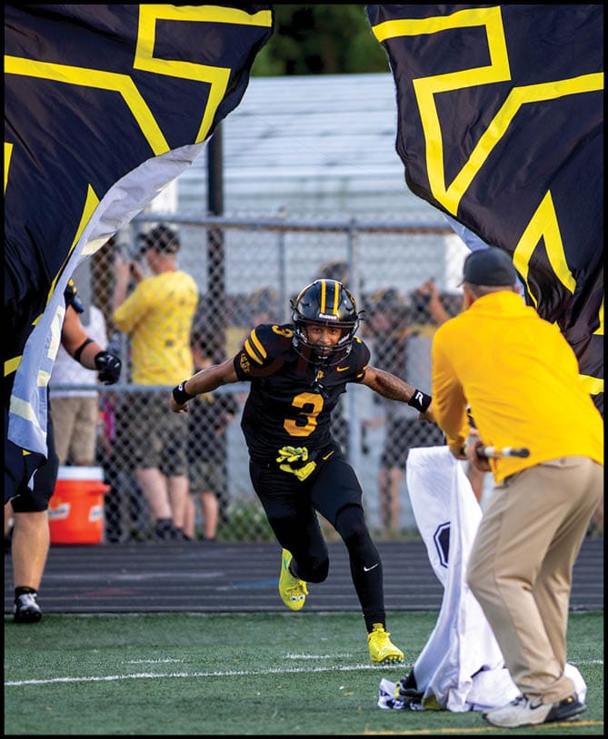 A person in a black and yellow football uniform runs through two large black flags with yellow designs held by two other people on a sports field. Description generated by AI