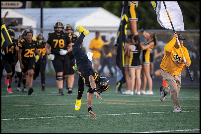 An adult in a black and yellow football uniform is performing a cartwheel on a football field while teammates and cheerleaders in similar colors watch and cheer in the background. Description generated by AI