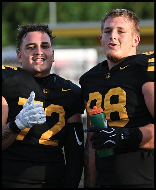 Two adults wearing black and yellow football uniforms are standing close together with one giving a thumbs up and the other holding a green water bottle. Description generated by AI