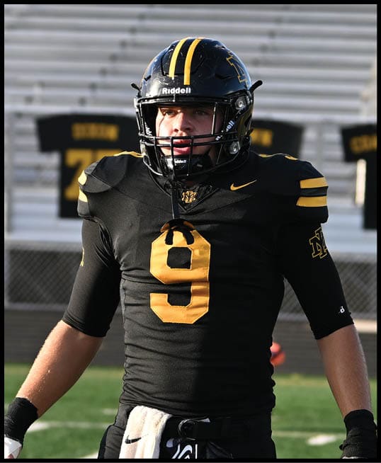 A person wearing a black sports uniform with the number nine in yellow is standing on a field with a white towel and black gloves attached to their waist. Description generated by AI