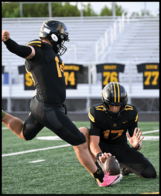 Two people in black and yellow football uniforms are on a field with one holding the ball on the ground while the other prepares to kick it. Description generated by AI