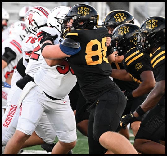 Four people in black and yellow football uniforms are lined up on a field preparing for a play while a person in a white and red uniform stands in the background. Description generated by AI