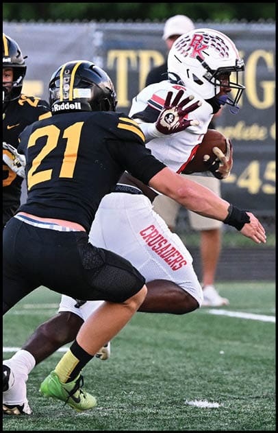 Two people in black and yellow football uniforms are attempting to tackle a person in a white and red football uniform who is holding a football and running on a field. Description generated by AI