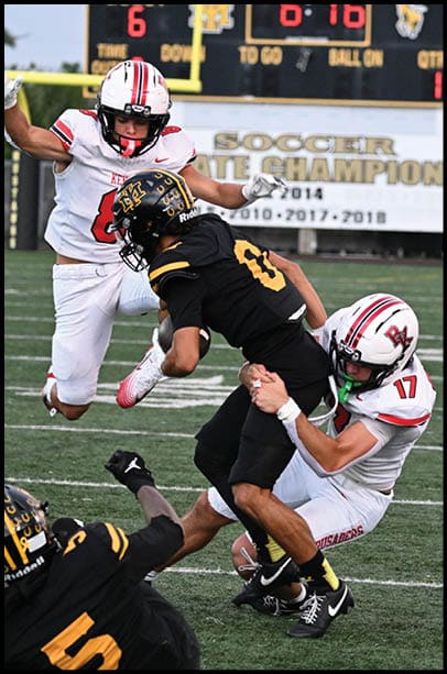 Four people in football uniforms are actively engaged in a play on a football field with a scoreboard and championship banner visible in the background. Description generated by AI
