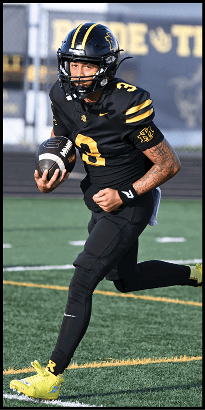 A person wearing a black football uniform with yellow stripes and bright yellow cleats is running on a green field. Description generated by AI