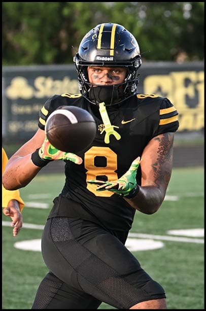 A person in a black football uniform with the number 8 is catching a football on a field while another person in a yellow hoodie stands nearby. Description generated by AI