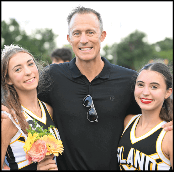A person in a black shirt stands smiling with arms around two people wearing cheerleading uniforms holding flowers and a sash, outdoors on a sports field. Description generated by AI