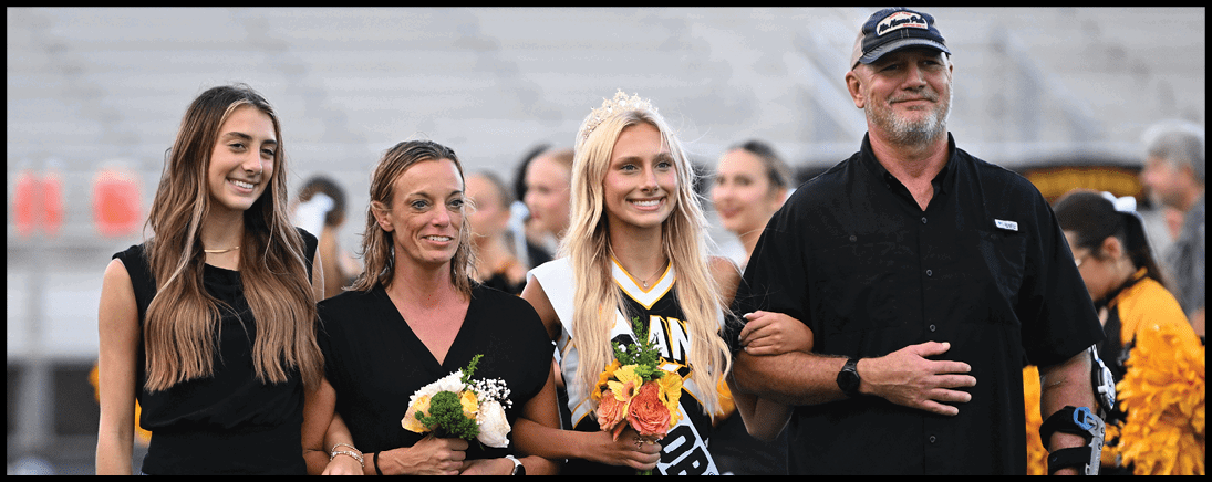 Four people stand arm in arm on a sports field, with one person wearing a cheerleading uniform and sash while holding flowers. Description generated by AI