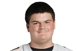 A person with short dark hair is wearing a light-colored sports jersey and smiling slightly against a plain white background. Description generated by AI