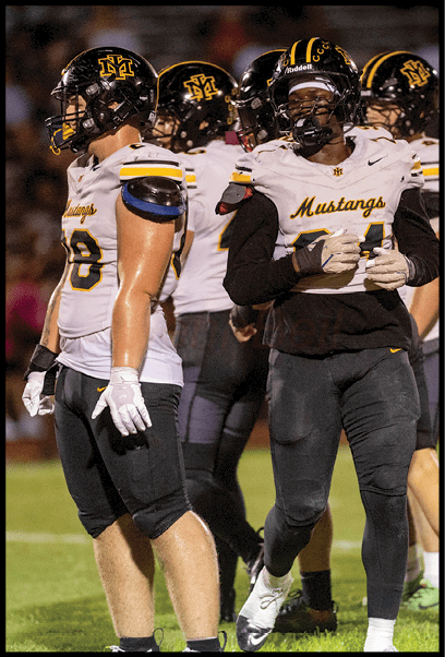 Four people wearing football uniforms with helmets stand on a grassy field during a game or practice. Description generated by AI