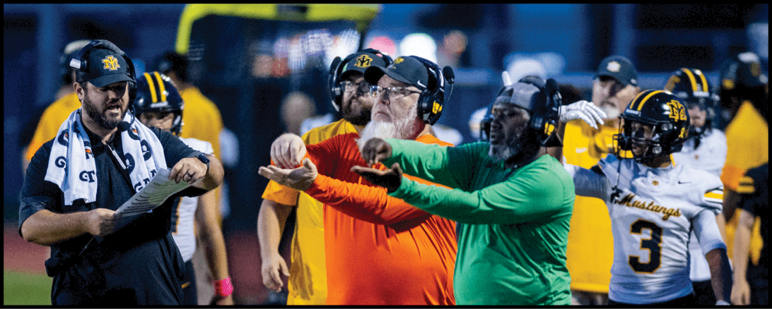 People wearing colorful shirts and pants are standing closely together on a grassy field, with one person holding a piece of paper. Description generated by AI