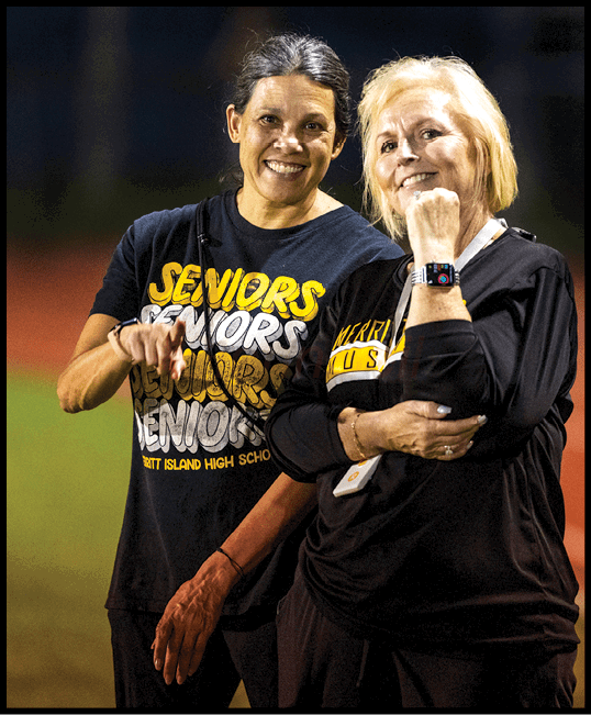 Two adults wearing black shirts with yellow and white text stand closely together, one pointing and the other showing a smartwatch. Description generated by AI