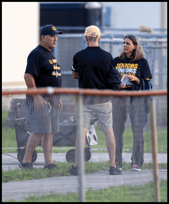 An adult with shoulder-length hair wearing a black shirt with yellow text stands outdoors near a fence holding a small item while another person stands nearby. Description generated by AI
