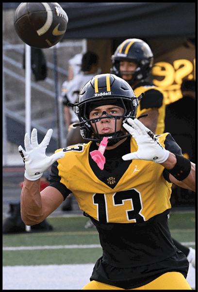 A person wearing a black and yellow football uniform with the number 13 and a helmet is reaching out with both hands to catch a football. Description generated by AI