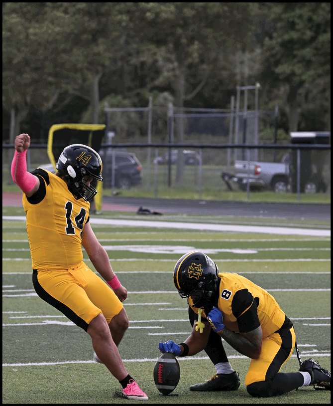 Two people in yellow and black football uniforms are on a field with one holding a football upright while the other prepares to kick it. Description generated by AI
