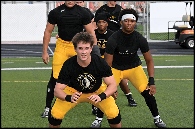 Four people wearing black shirts and yellow pants are positioned on a sports field, with one person crouching in front and the others standing behind. Description generated by AI