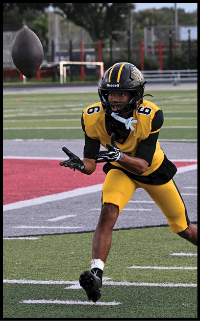 A person wearing a yellow and black football uniform with the number six is reaching out with both hands as if to catch a ball on a football field. Description generated by AI