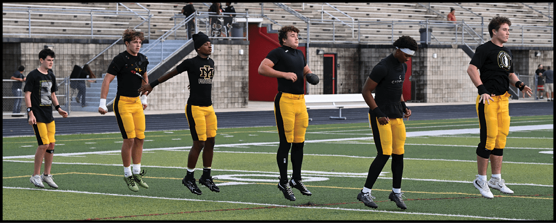 Four adults wearing black shirts and yellow pants are standing on a sports field with bleachers in the background. Description generated by AI