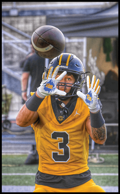 A person wearing a yellow and blue football uniform with the number three and a helmet is reaching up with a gloved hand to catch a football in midair. Description generated by AI