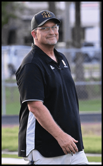 A person wearing glasses, a black cap, and a black sports shirt stands outdoors on a grassy field with a slight smile. Description generated by AI