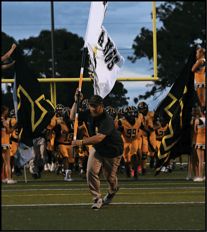 A person holding a white flag runs ahead of a group of football players in yellow uniforms and cheerleaders in yellow and white outfits on a football field at dusk. Description generated by AI