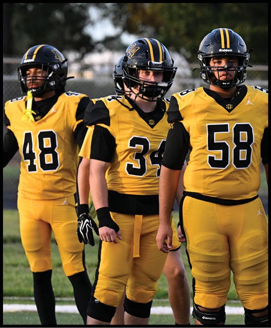 Four people wearing yellow and black football uniforms stand on a field, appearing to prepare for a game or practice. Description generated by AI