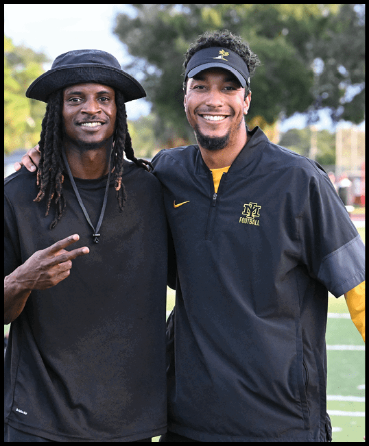 Two people stand closely together outdoors on a sports field, smiling with one person making a peace sign and both wearing casual athletic clothing including a hat and a visor. Description generated by AI