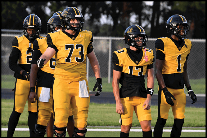 Five people wearing yellow and black football uniforms stand on a grassy field with a chain-link fence in the background. Description generated by AI