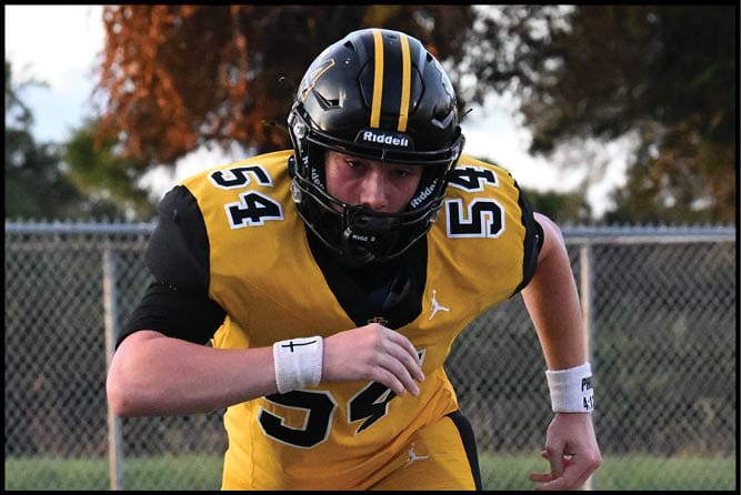 A person wearing a yellow and black football uniform with the number 54 is running on a track field with a chain-link fence and trees in the background. Description generated by AI