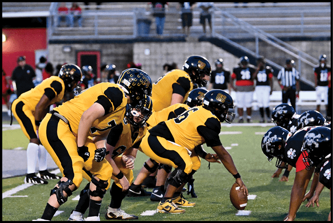 A group of people in yellow and black football uniforms are lined up on a field preparing for a play with others and spectators visible in the background. Description generated by AI