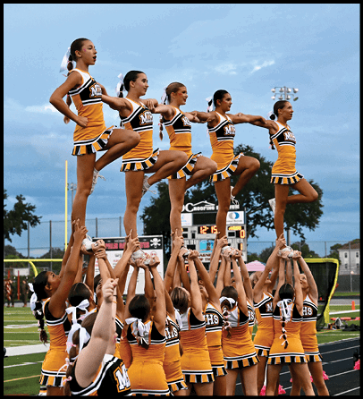 People in yellow and black cheerleading uniforms are performing a stunt with their legs raised and supported by others' hands against a background of a scoreboard and trees. Description generated by AI