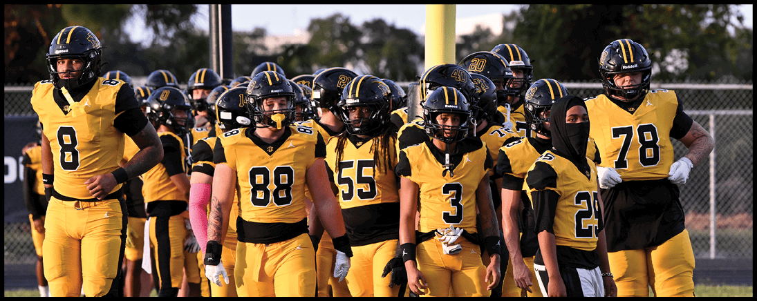A group of people wearing yellow and black football uniforms stand together on a field with one person in white pants among them. Description generated by AI