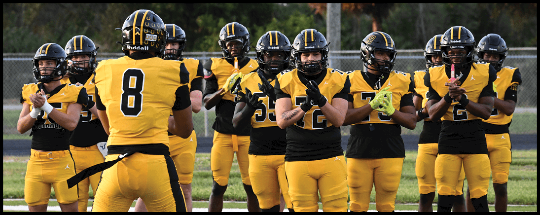 A group of people in yellow and black football uniforms stand in a line on a field, clapping and facing a person wearing a jersey with the number 8. Description generated by AI