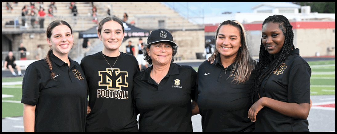 Five adults wearing black sports apparel stand closely together smiling on a football field with bleachers and other people in the background. Description generated by AI