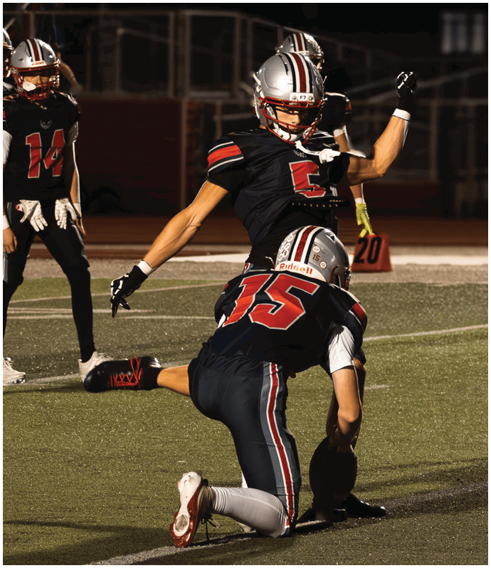Two people in football uniforms are on a field with one holding the ball on the ground while the other prepares to kick it. Descripci n generada con IA