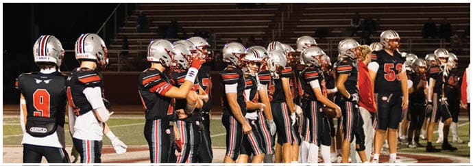 A group of people wearing football helmets and uniforms stand in a line on a field with empty bleachers in the background at night. Descripci n generada con IA