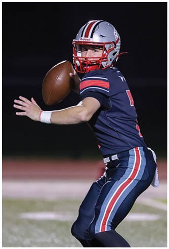 A person wearing a football helmet and uniform is preparing to throw a football against a dark background. Descripci n generada con IA
